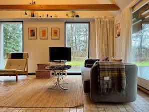 Ein Wohnzimmer mit Fernseher, einer Couch, einem Sessel, einem Tisch und einem großen Fenster mit Blick auf Bäume.