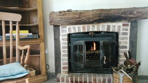 A fireplace with a brick surround and a wooden mantel, a wooden chair with a cushion, and a bookshelf with books. - Cottage in château grounds near Rouen (Limésy)