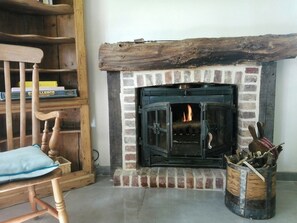 A fireplace with a brick surround and a wooden mantel, a wooden chair with a cushion, and a bookshelf with books. - Cottage in château grounds near Rouen (Limésy)