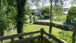 A wooden balcony railing overlooking a lush garden with a stone wall and a road in the background. - Cottage in château grounds near Rouen (Limésy)