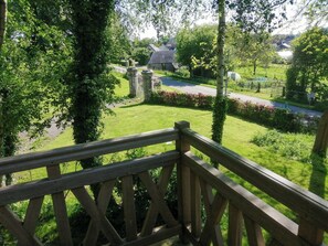 A wooden balcony railing overlooking a lush garden with a stone wall and a road in the background. - Cottage in château grounds near Rouen (Limésy)