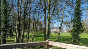 A wooden deck overlooking a well-maintained lawn with trees and a large building in the background. - Cottage in château grounds near Rouen (Limésy)