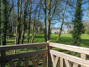 A wooden deck overlooking a well-maintained lawn with trees and a large building in the background. - Cottage in château grounds near Rouen (Limésy)