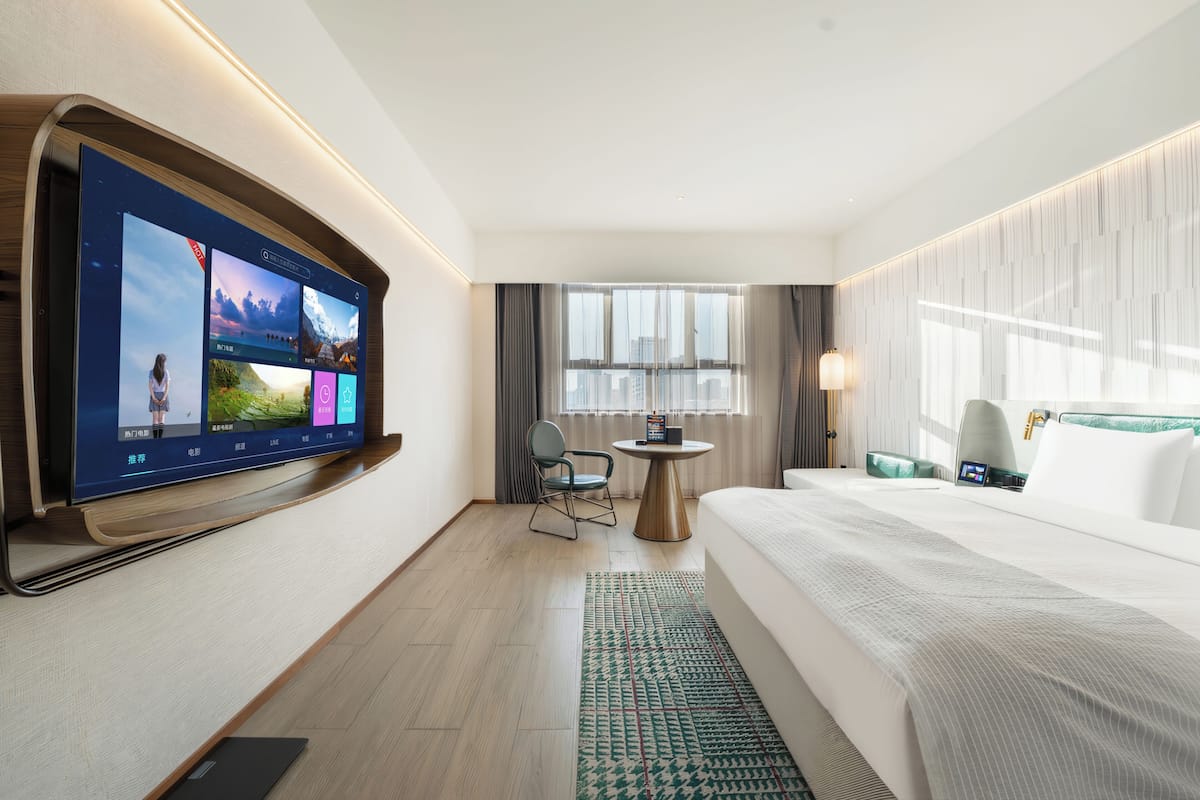 Habitación de hotel con un televisor de pantalla plana grande montado en la pared, una cama, una mesita redonda pequeña, una silla y una ventana con cortinas.