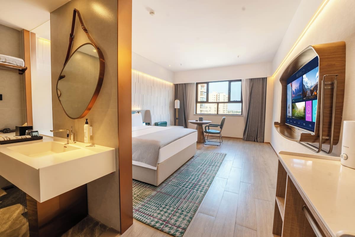 Habitación de hotel con una cama grande, un escritorio, un televisor y un baño con un espejo grande y lavamanos.
