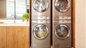 A laundry area with two stacked washing machines, a wooden cabinet, a sink, and a window with blinds.