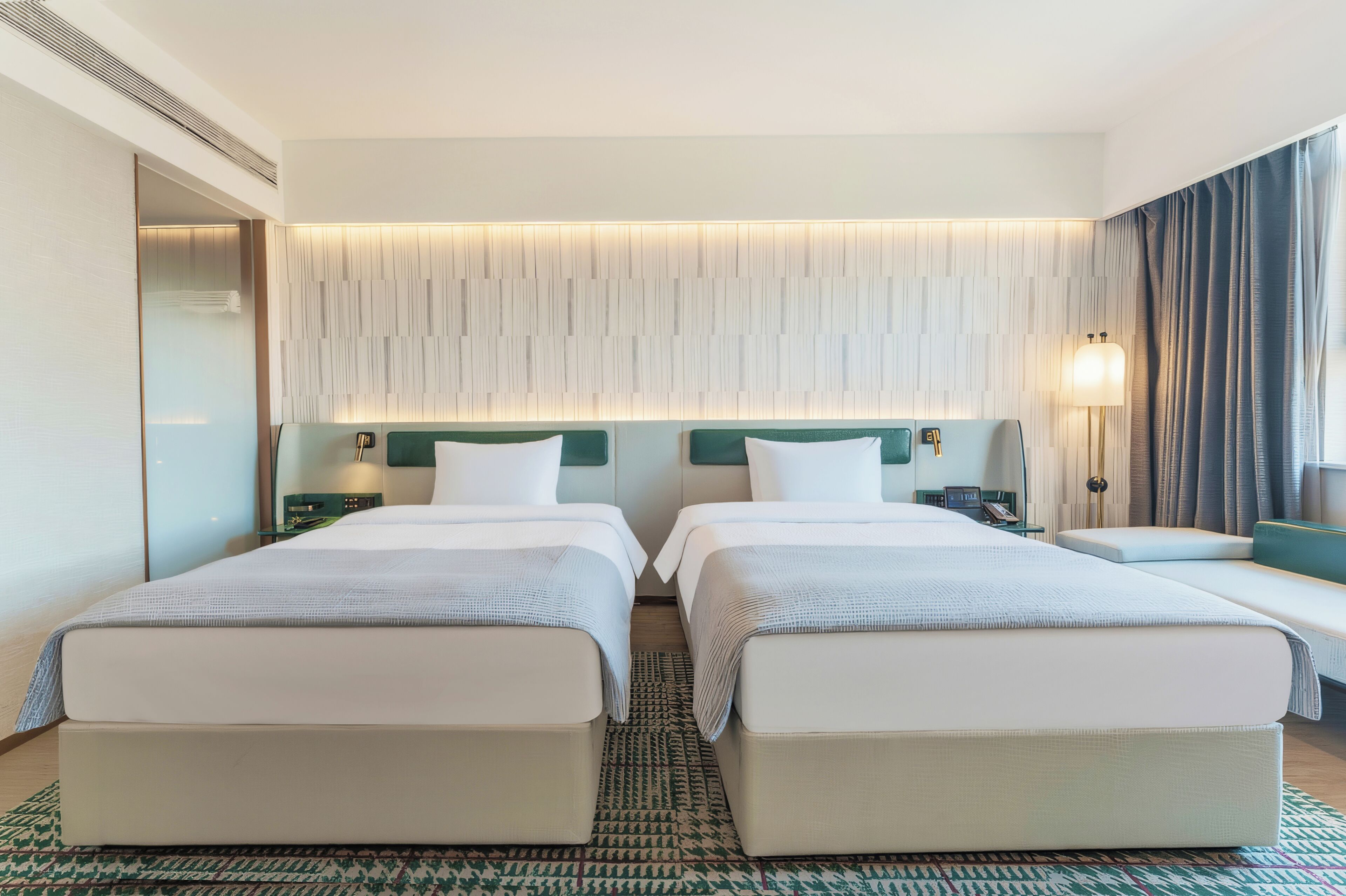 Ein Hotelzimmer mit zwei Betten, einem Nachttisch mit Lampe, einem Spiegel und einem gemusterten Teppich.