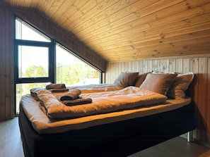 A loft-style bedroom with a sloped ceiling, a large bed with a quilted blanket, and a view of trees through a window.