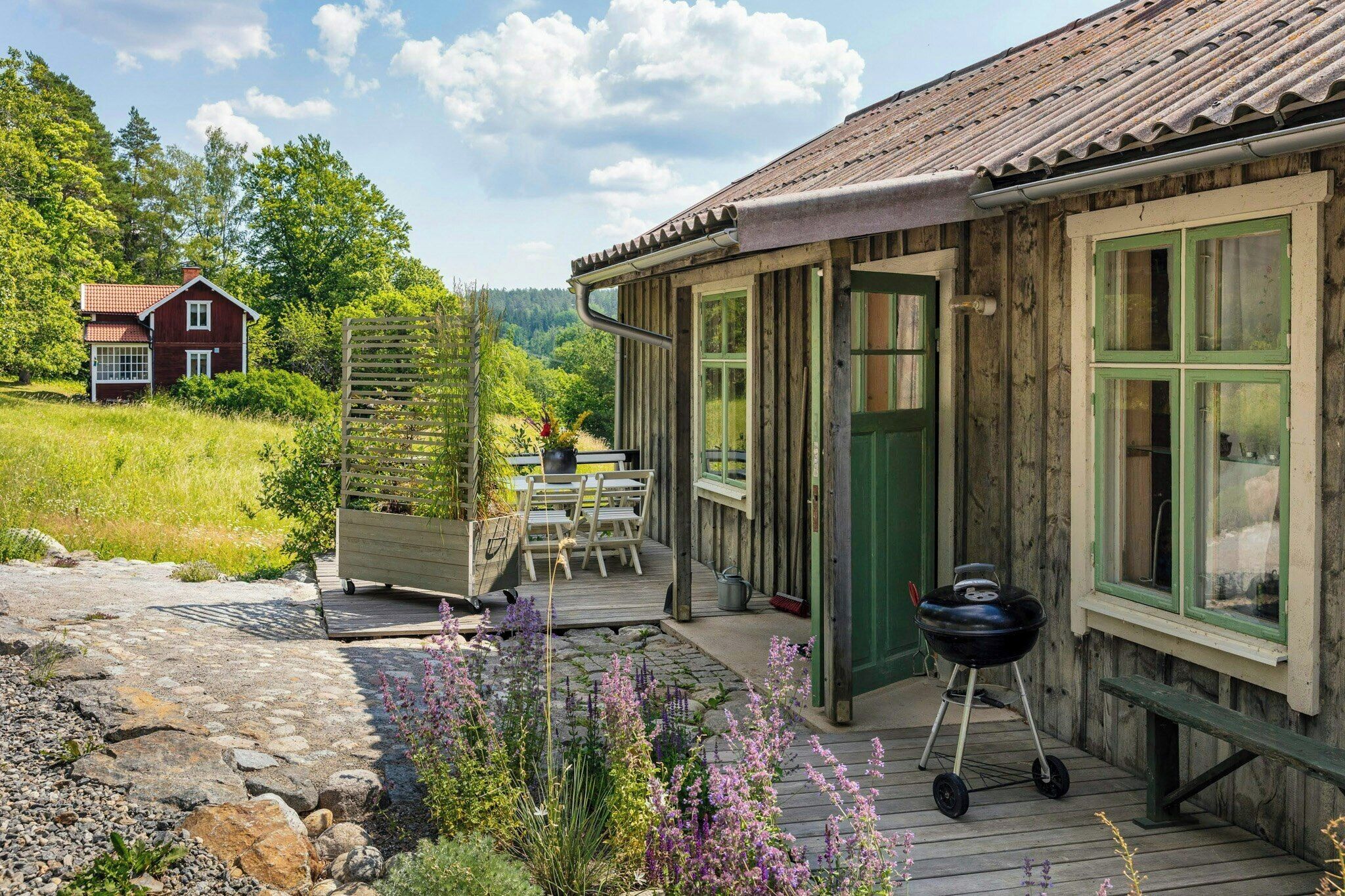 A wooden cabin with a deck, a barbecue grill, and a view of a red house in the background.