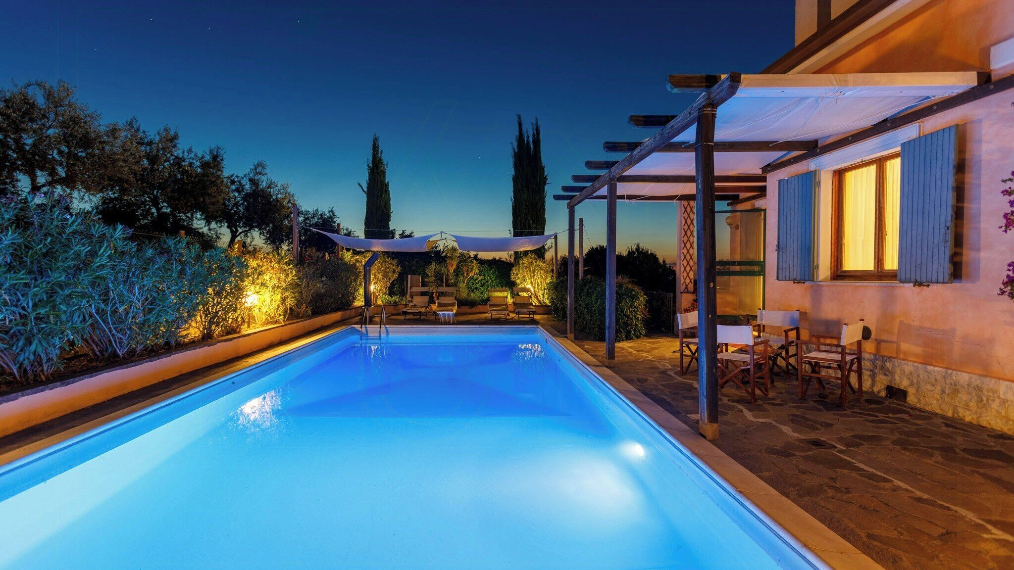 A swimming pool with lounge chairs and a pergola, surrounded by vegetation and a building with shutters.