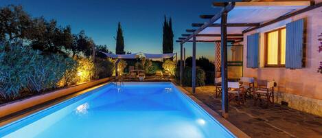 A swimming pool with lounge chairs and a pergola, surrounded by vegetation and a building with shutters.