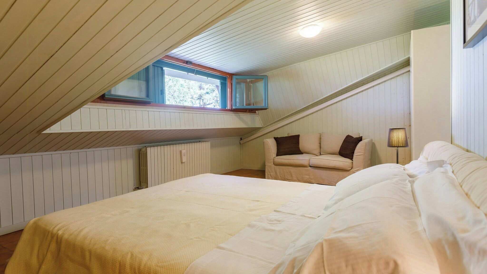 A bedroom with a sloped ceiling, a bed, a sofa, and a window.