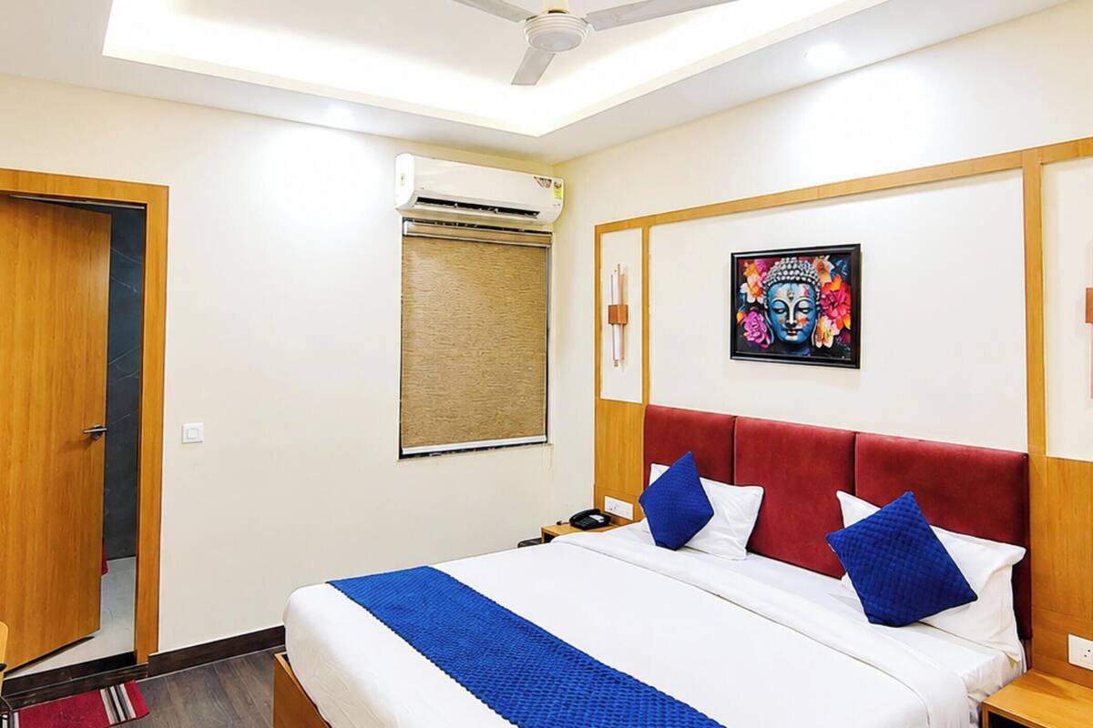 Una habitación de hotel con una cama, ventilador de techo, equipo de aire acondicionado, puerta de madera, una obra de arte colgada en la pared y un cabecero rojo.