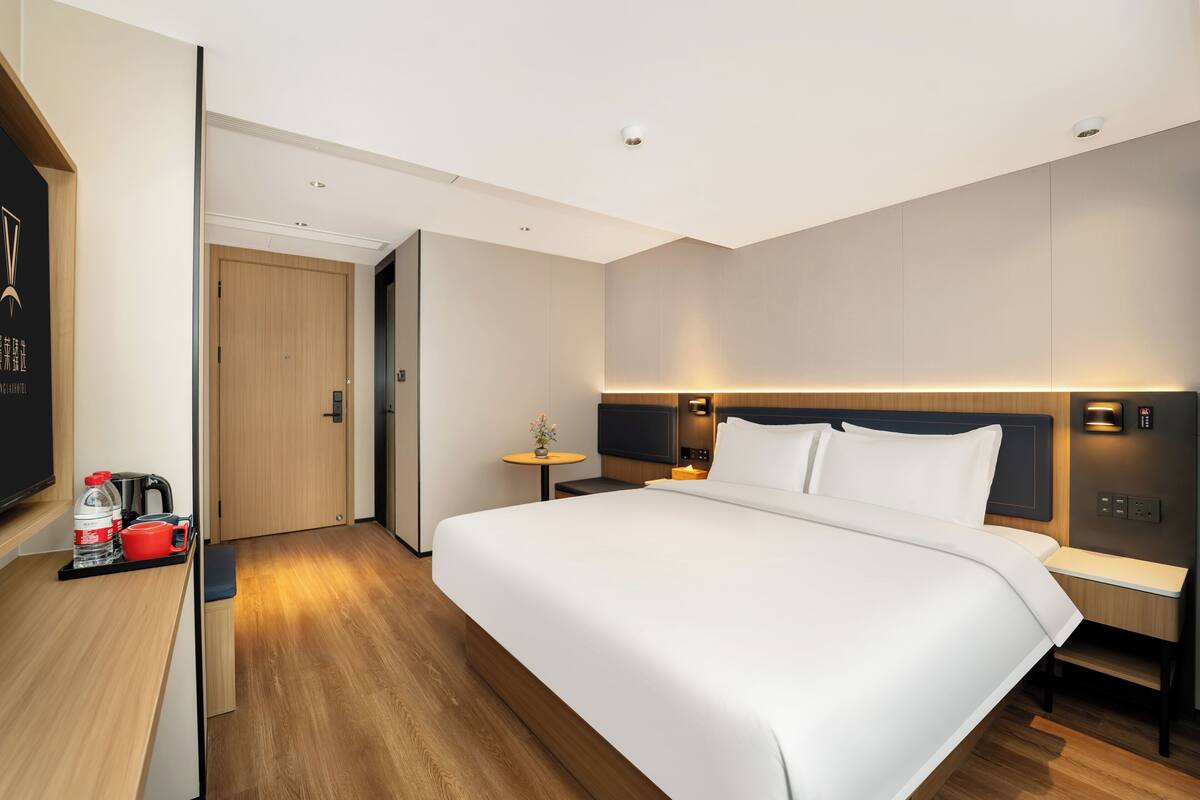Habitación de hotel con una cama grande, mesitas de noche, un televisor, una pequeña mesa con una planta y piso de madera.