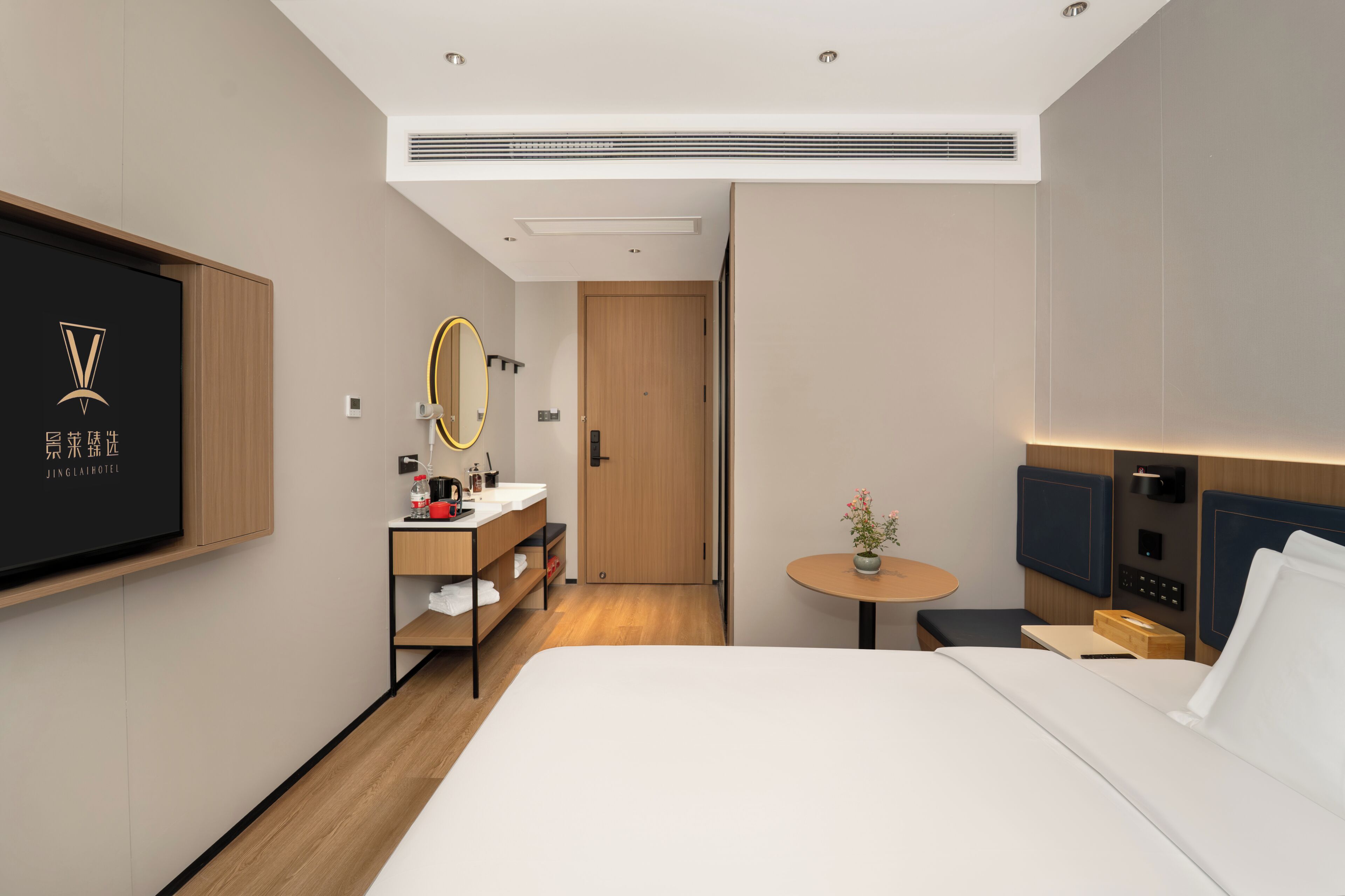 Ett modernt hotellrum med en stor säng, ett sängbord, en rund spegel, ett litet runt bord med en vas, en trädörr och en tv som är monterad på väggen.