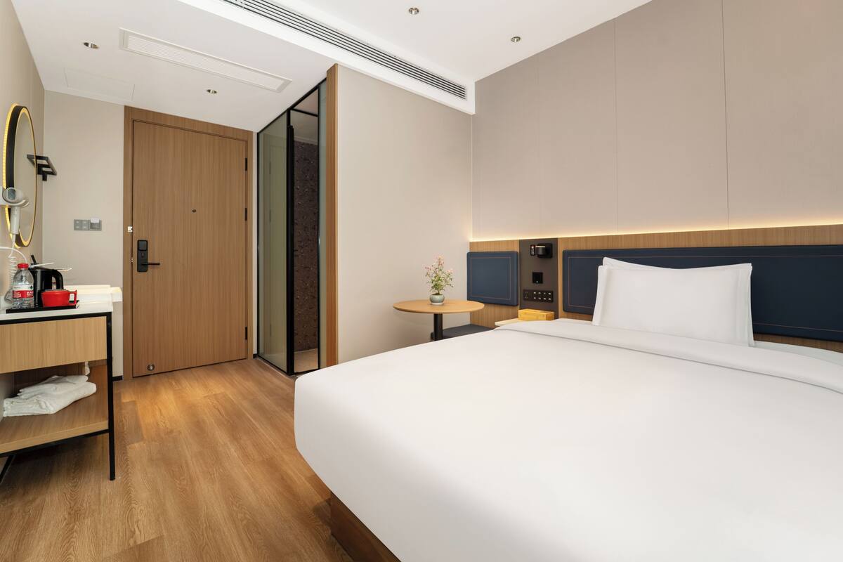 Habitación de hotel con una cama grande, un escritorio con cafetera, una mesita con planta y una puerta corredera.