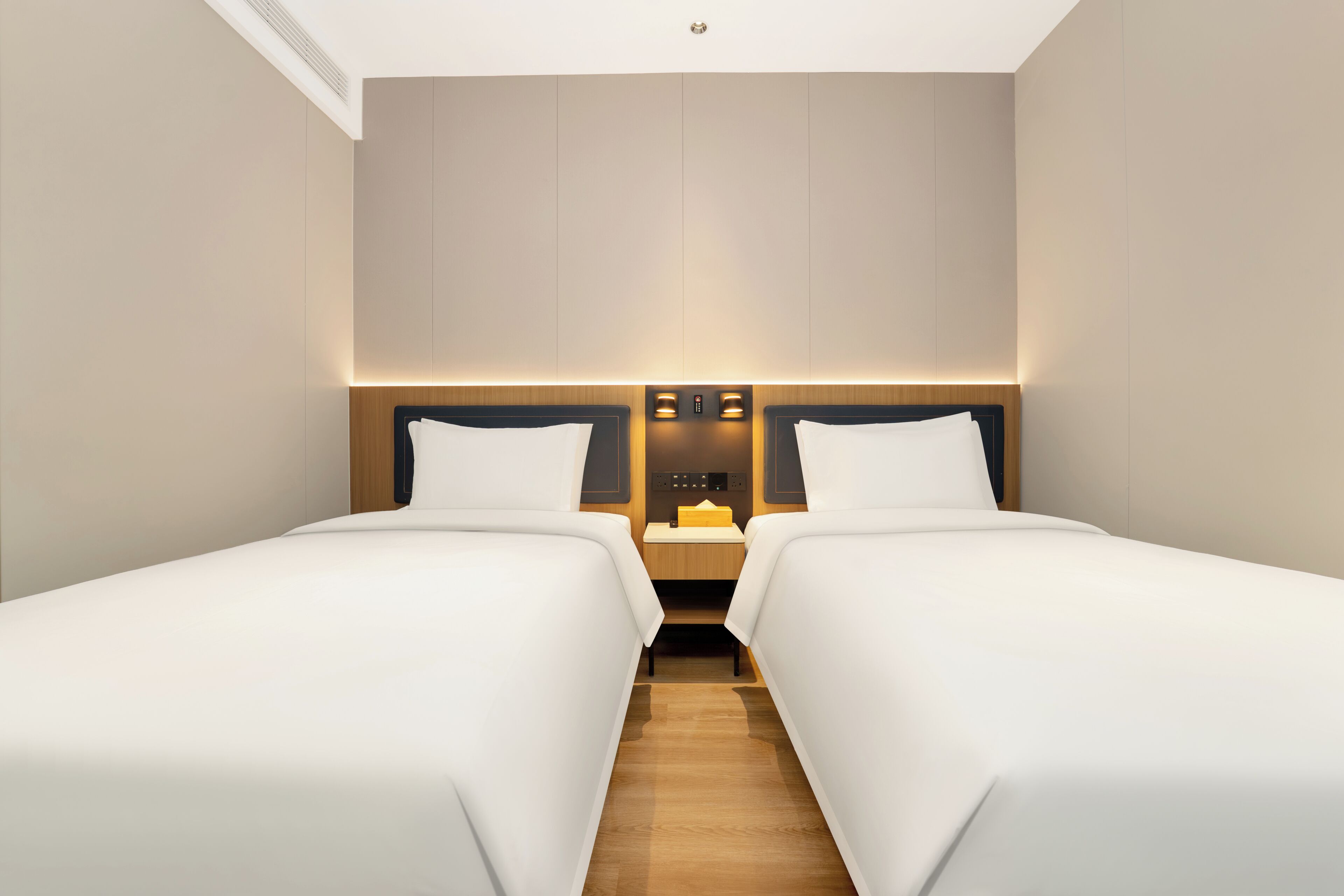 A hotel room with two beds, a wooden floor, a nightstand with a lamp, and a wall-mounted control panel.