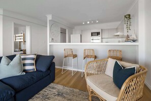 Ein modernes Wohnzimmer mit einer Couch, zwei Korbstühlen, einem Essbereich und einer Küche im Hintergrund sowie einer Uhr an der Wand.