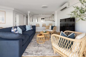 Ein modernes Wohnzimmer mit einer Couch, Korbstühlen, einem Couchtisch und einer Küche im Hintergrund.