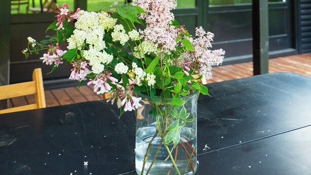 Et bord med en klar glasvase med blomster, en træstol og udsigt til et grønt udendørs område gennem store vinduer.