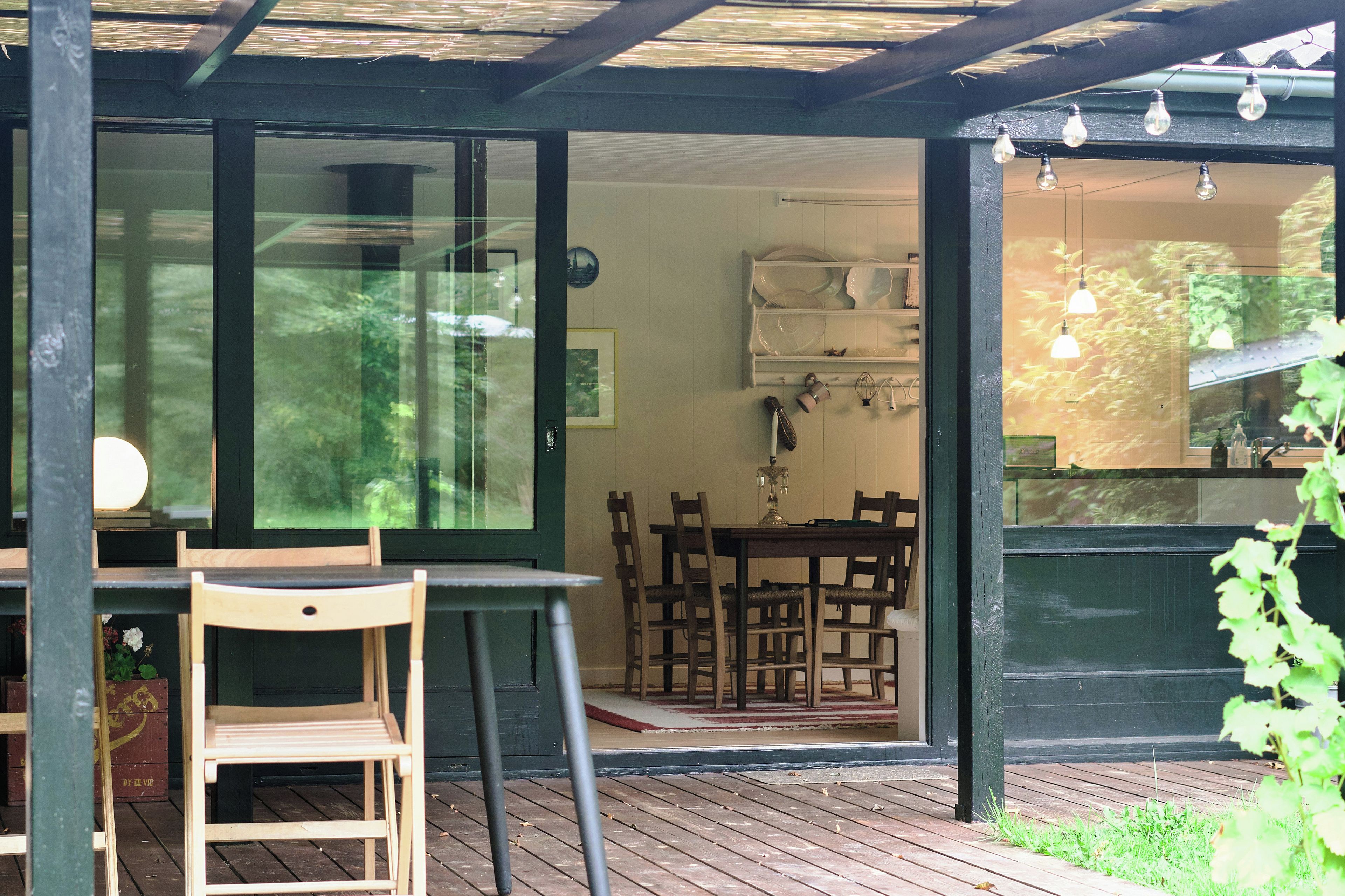 Een houten terras met een tafel en stoelen, een glazen deur naar een binnenruimte met een eettafel en stoelen, en een plank met decoratieve objecten.