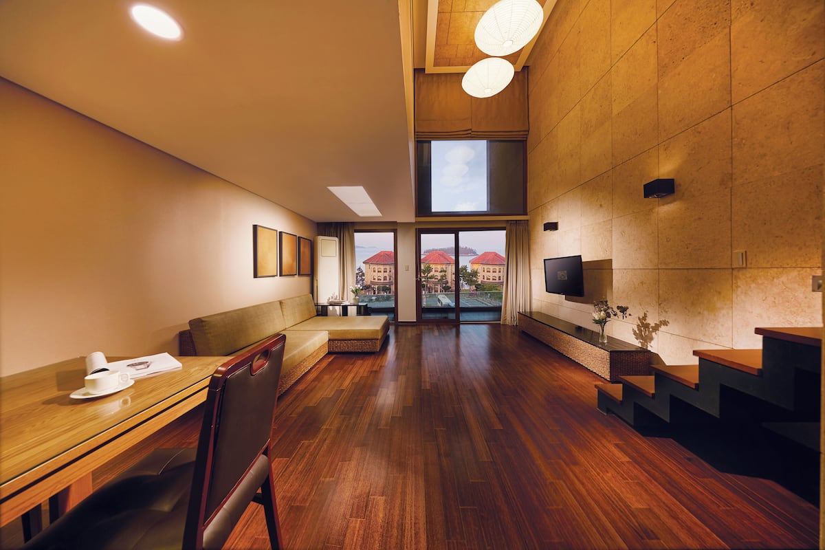 A spacious hotel lobby with a wooden floor, a sofa, a dining table, and a staircase.