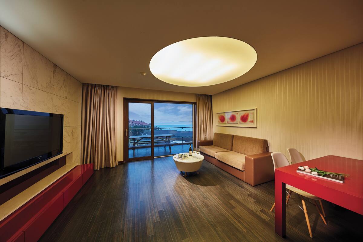 A hotel room with a television, a dining table, a sofa, a coffee table, and a large window offering a view.