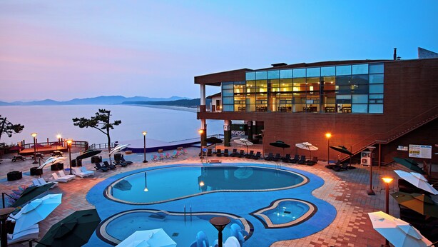 Un área de piscina con sillones de playa y sombrillas, un edificio con grandes ventanales y vistas al agua.