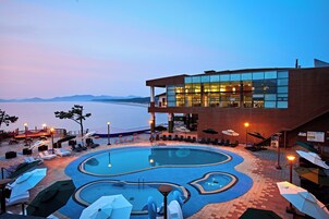 Un área de piscina con sillones de playa y sombrillas, un edificio con grandes ventanales y vistas al agua.