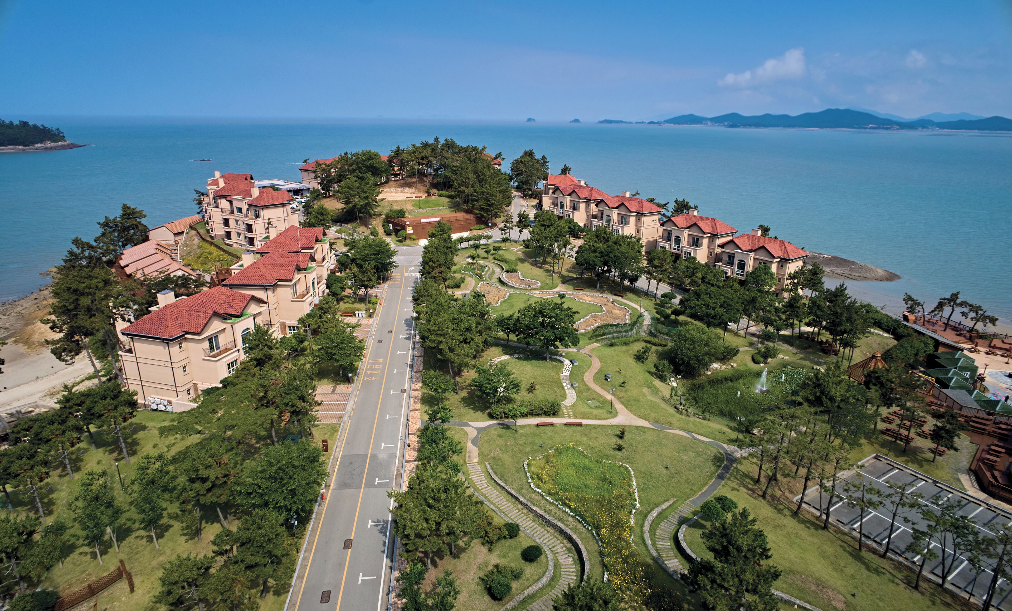 A coastal residential area with a central road, green spaces, and buildings with red roofs.