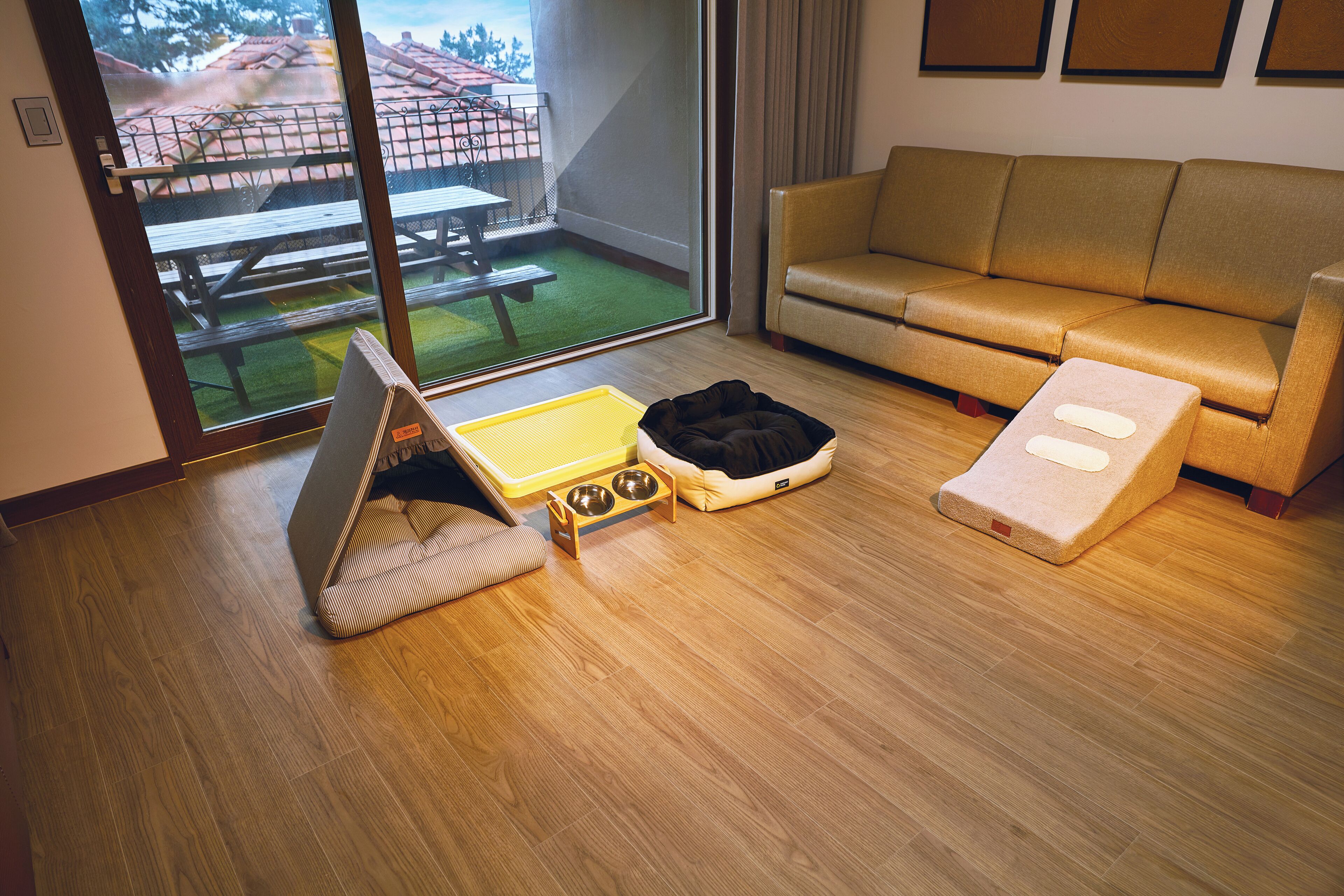 Ein Zimmer mit einer Couch, einem Haustierbett, einer Futterstelle für Haustiere und einem Fenster mit Blick auf eine Terrasse.