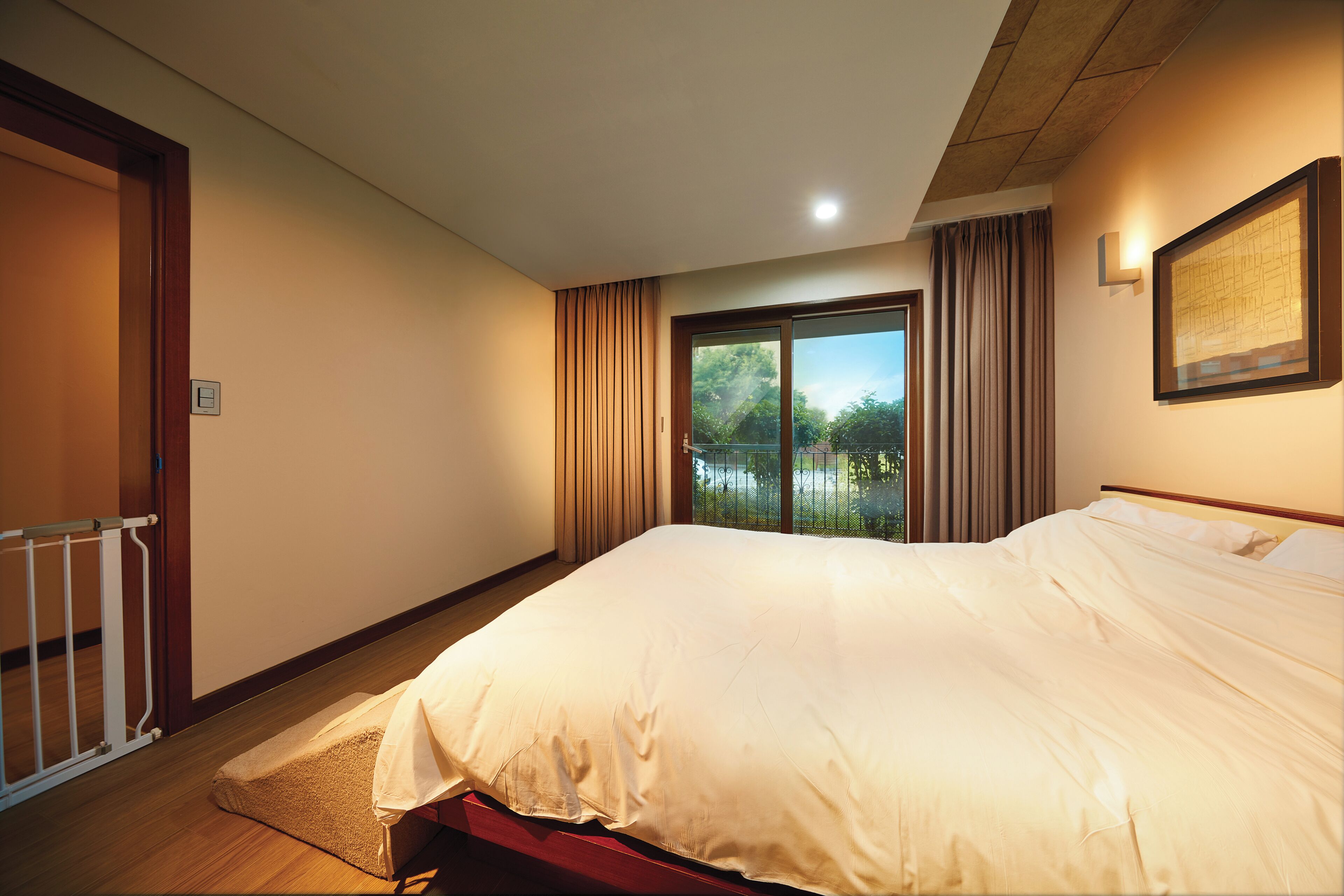 A bedroom with a large bed, a window with curtains, a door, and a framed picture on the wall.