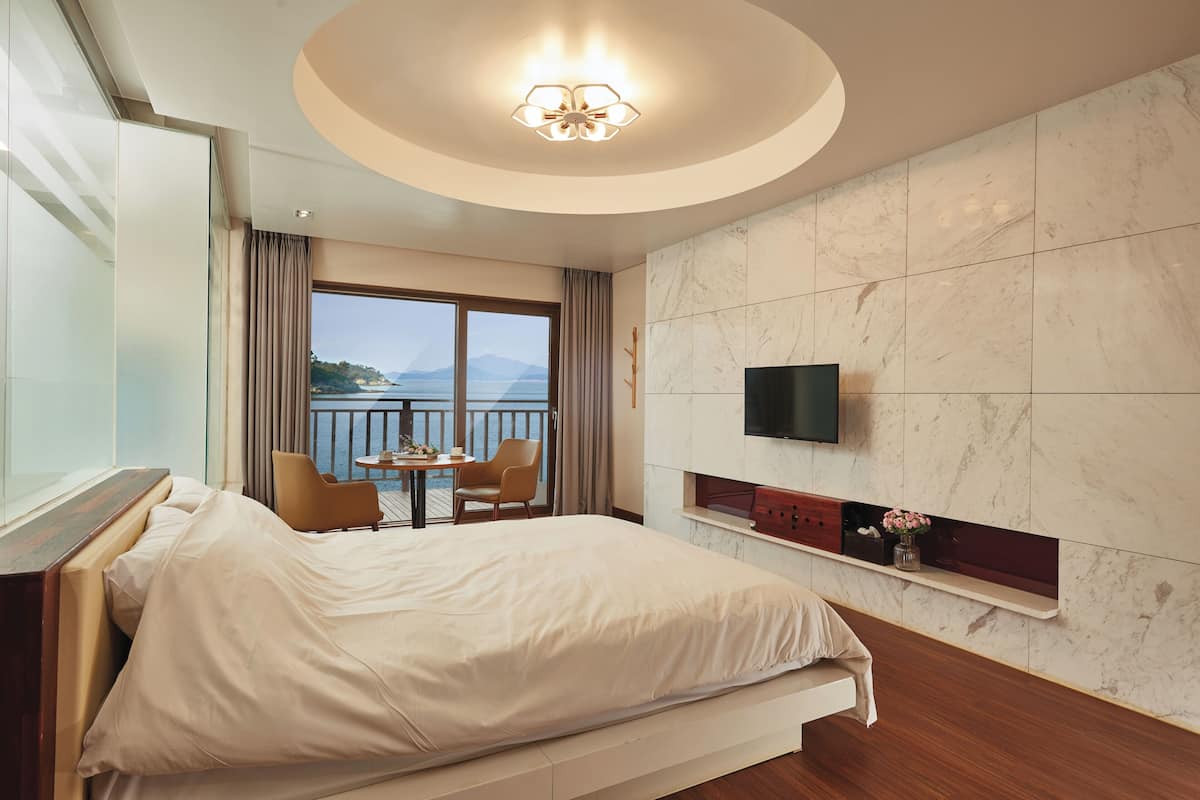 A hotel room with a bed, a TV, a small table with chairs, and a balcony with a view of the sea.