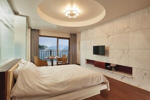 Habitación de hotel con cama, televisión, una pequeña mesa con sillas y un balcón con vista al mar.