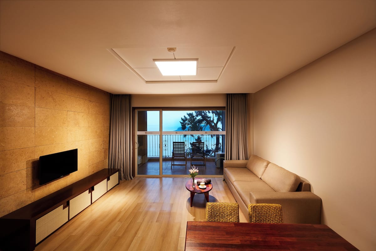 A modern living room with a flat-screen TV, wooden flooring, a sofa, a coffee table, and a balcony with a view.