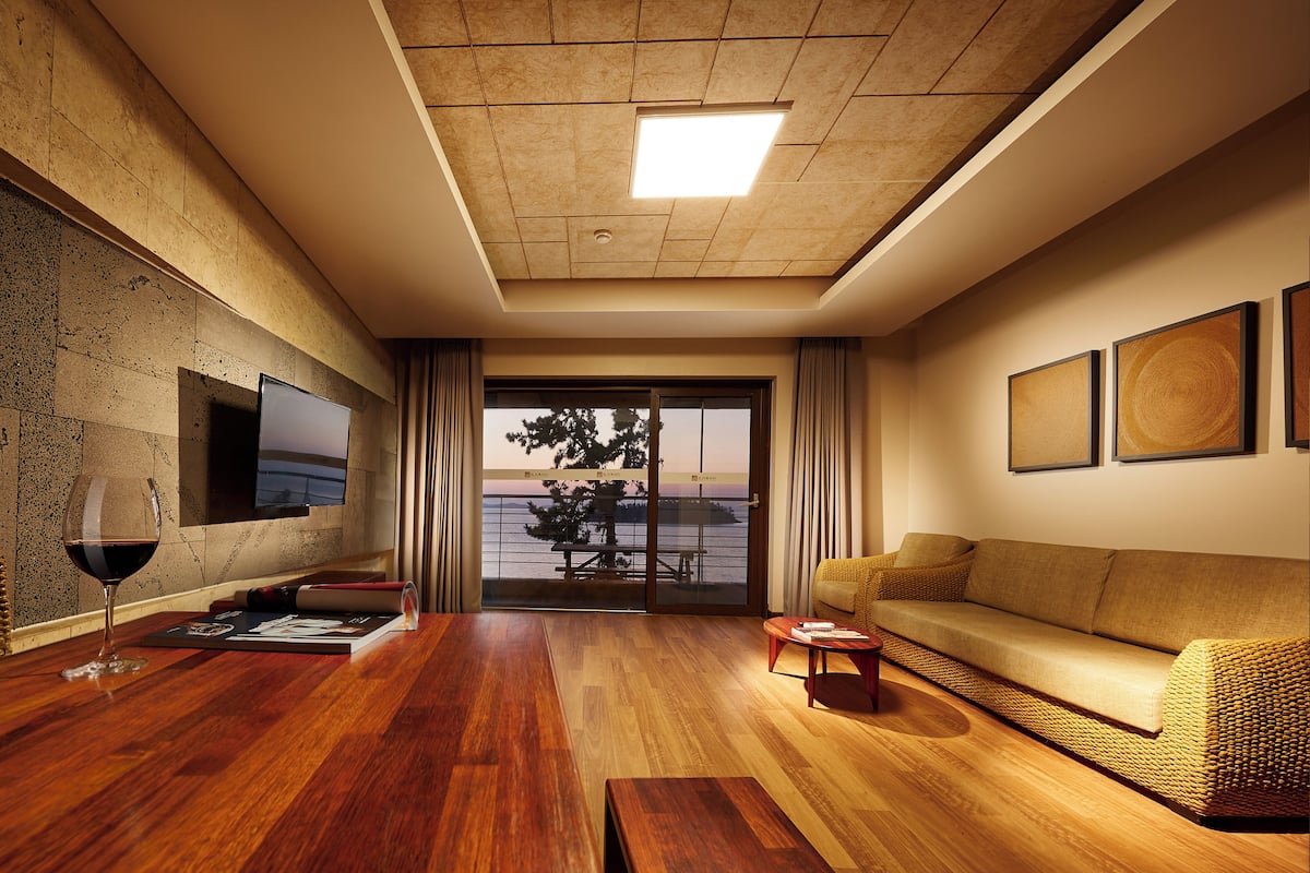 A modern living room with a wooden floor, a sofa, a glass door leading to a balcony, and a flat-screen TV mounted on the wall.