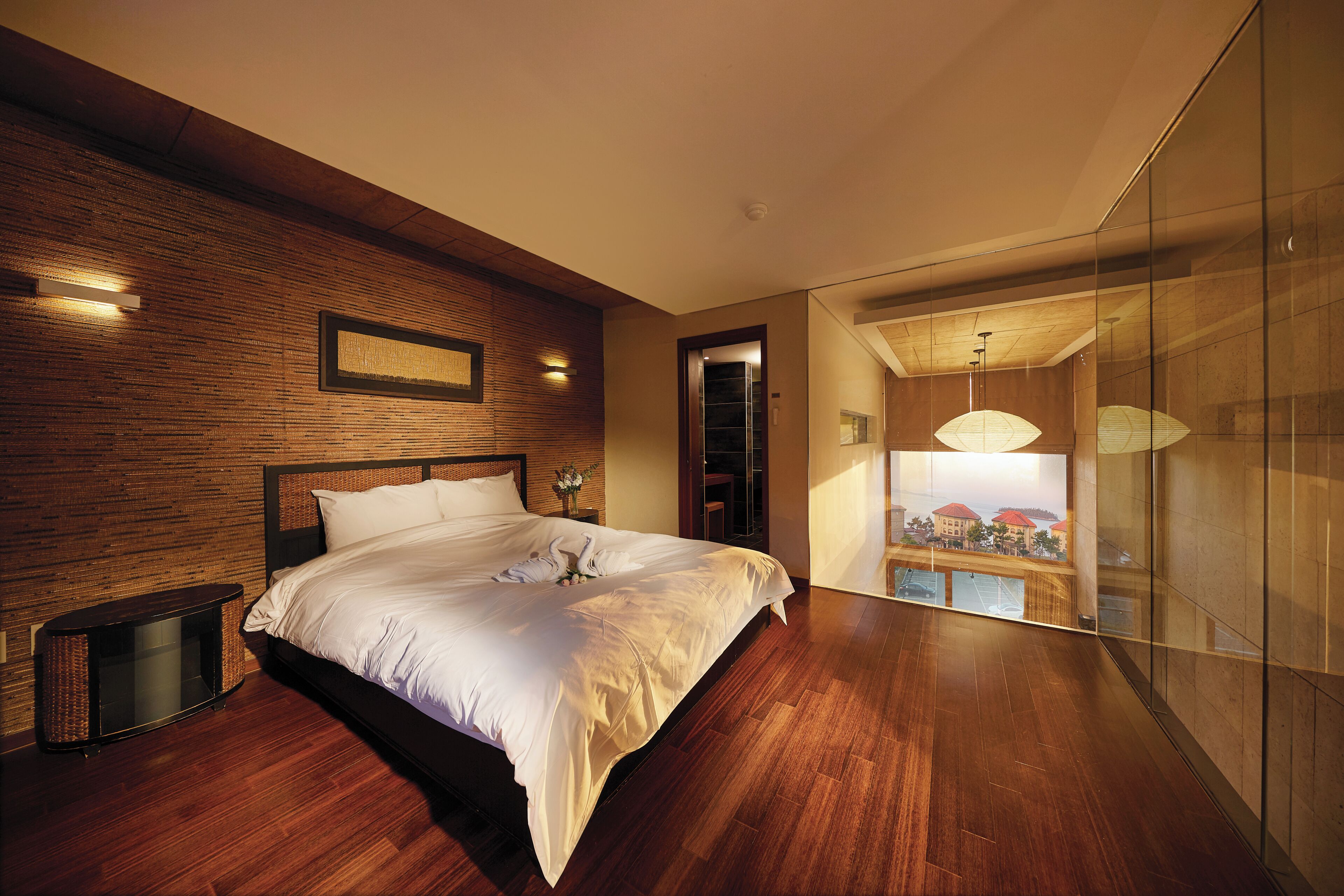 Un dormitorio moderno con piso de madera, una cama grande, televisor montado en la pared y un separador de vidrio con vistas.