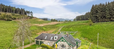 Una casa de piedra con tejado verde, rodeada de campos y árboles verdes, bajo un cielo despejado.