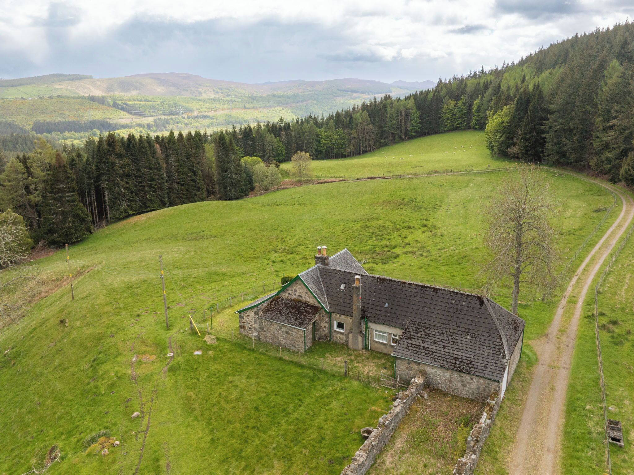 Een landhuis met een stenen gevel, omgeven door groene velden en een beboste heuvel.