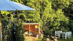 A wooden deck with a covered seating area, surrounded by lush greenery and white chairs.