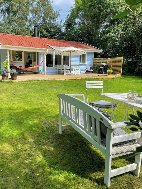Une maison avec un toit rouge, une terrasse couverte meublée de mobilier blanc et une cour verdoyante.