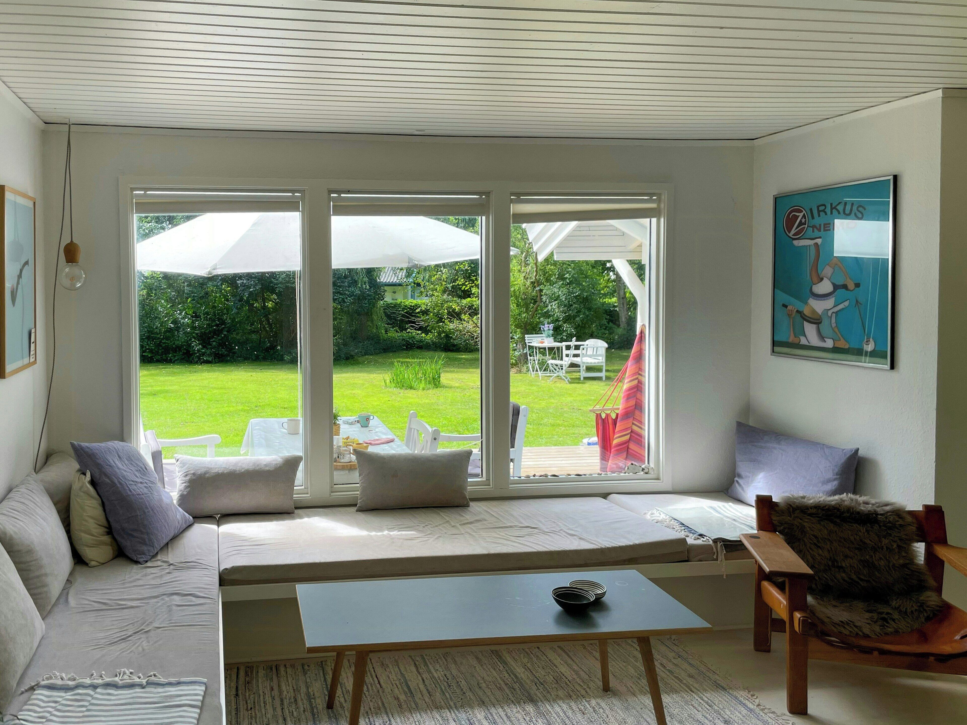 A room with a sofa, a small table, and a chair. There is a large window with a view of a garden and a patio.