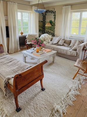 A cozy living room with a sofa, a coffee table with books and a drink, a wooden bench, and a large window with curtains. - Charming farmhouse with open spaces near Österlen (Löderup)