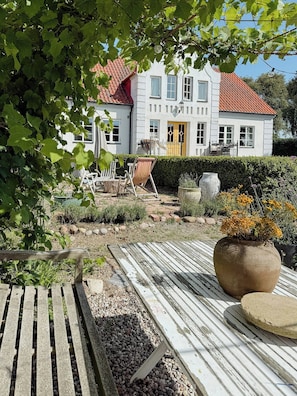 A white building with a red roof, a wooden deck, and a garden with plants and a large pot. - Charming farmhouse with open spaces near Österlen (Löderup)