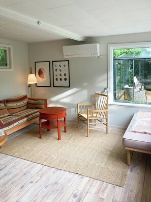 Ein Zimmer mit einer Couch, einem runden Tisch, einem Stuhl, einer Lampe und einem Fenster mit Blick ins Grüne.