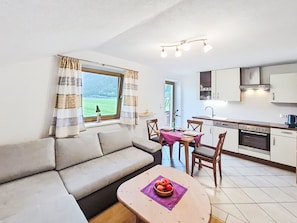 Ein Wohnzimmer mit einer Couch, einem Esstisch mit Stühlen, einer Küchenzeile mit Schränken und Geräten sowie einem Fenster mit Vorhängen.
