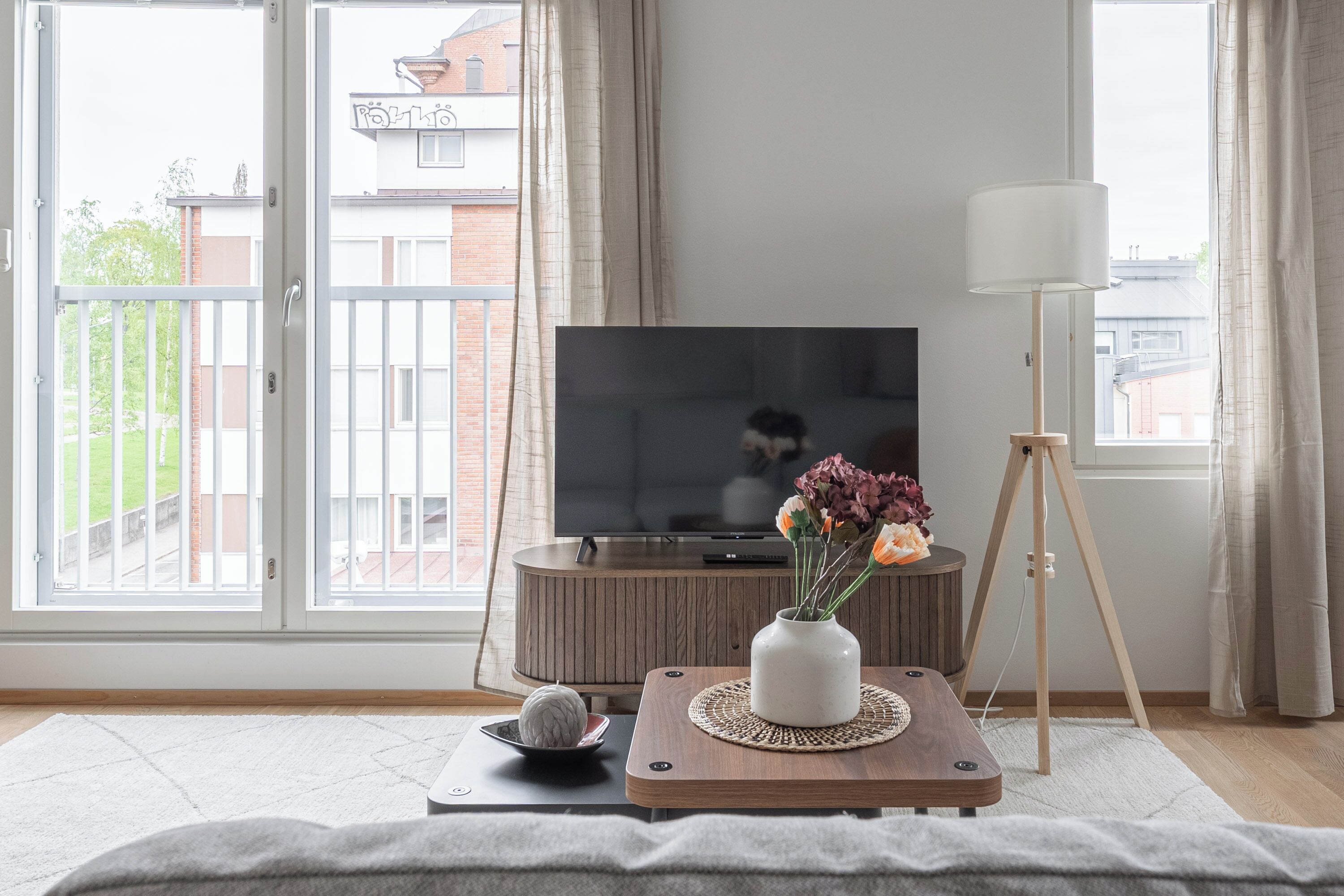 A living room with a TV, a wooden coffee table with a vase of flowers, a lamp, and a view of buildings through large windows.