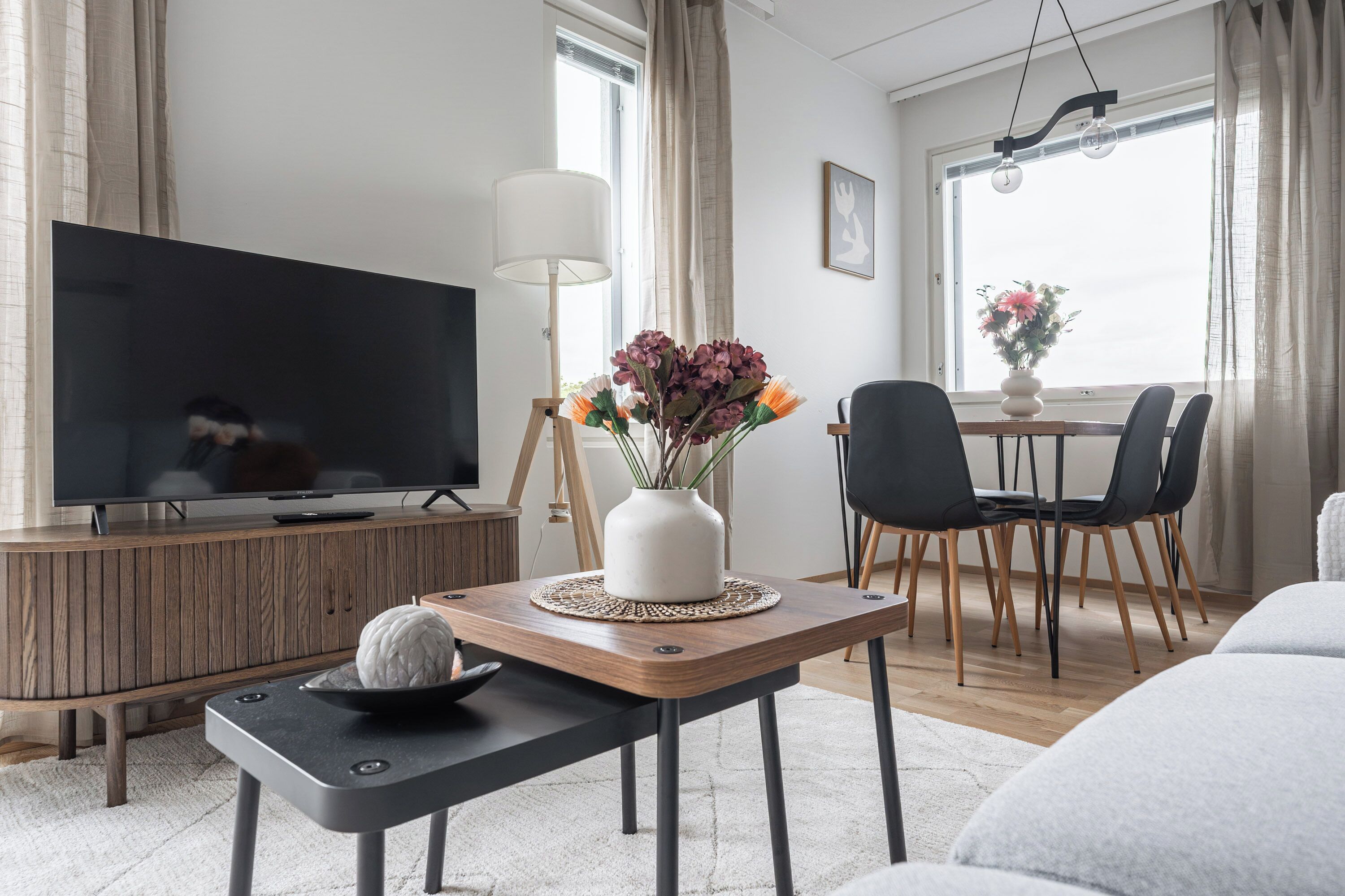 Una sala de estar moderna con un televisor de pantalla plana, una pequeña mesa con un jarrón decorativo, un rincón de comedor con sillas y un sofá.