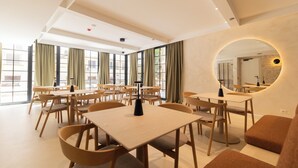 Un espace repas moderne doté de plusieurs tables et chaises, de grandes fenêtres et d’un grand miroir rond au mur.