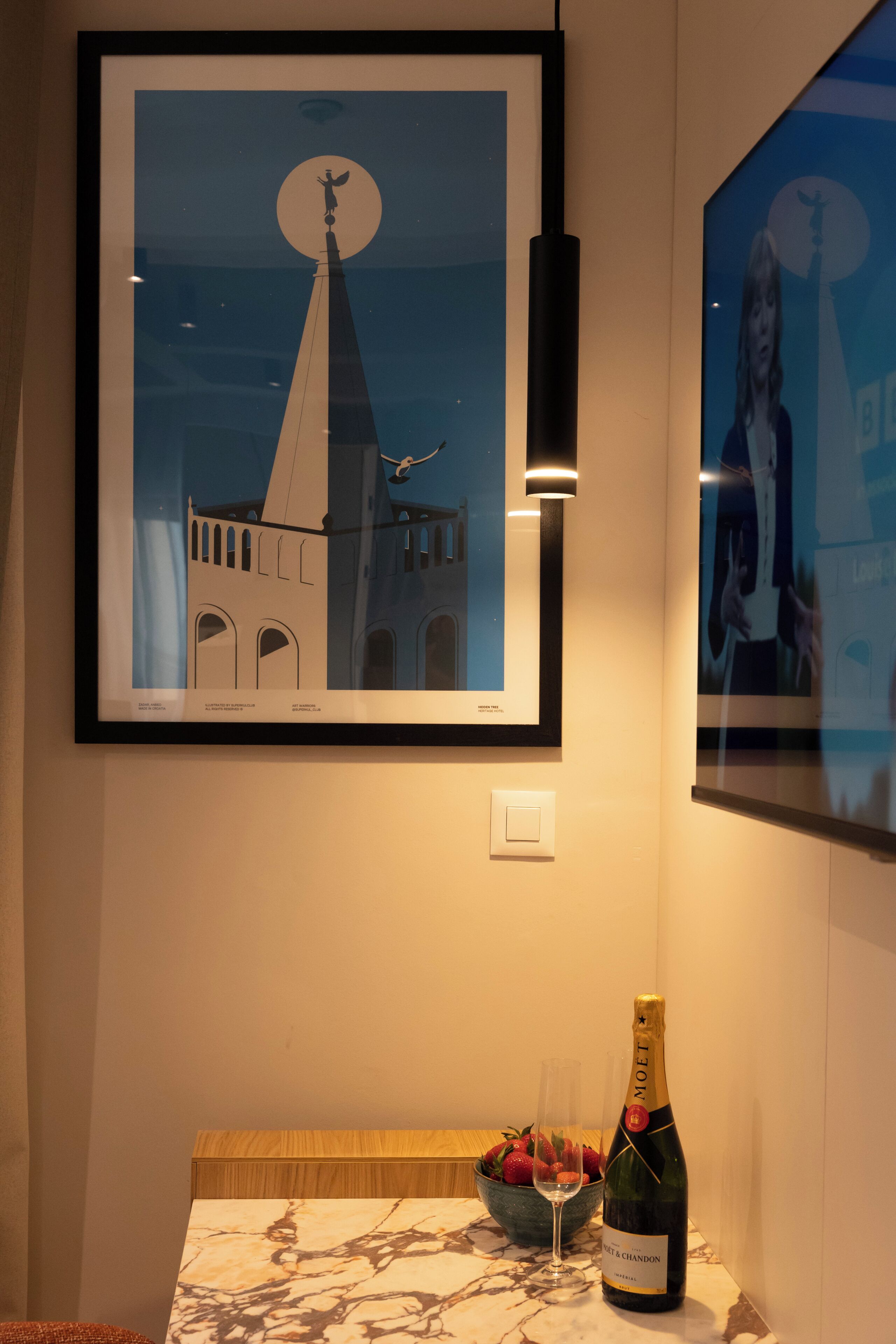 A framed artwork of a building with a tower, a bottle of champagne, and a bowl of strawberries on a table.